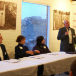 People seated at a long table while a man speaks into a microphone